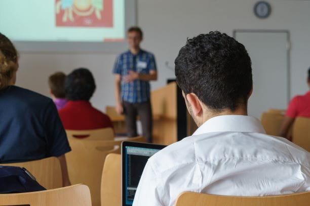 Mann in weißem Hemd sitzt in Publikum mit Laptop in einer Rede auf einer Tagung.